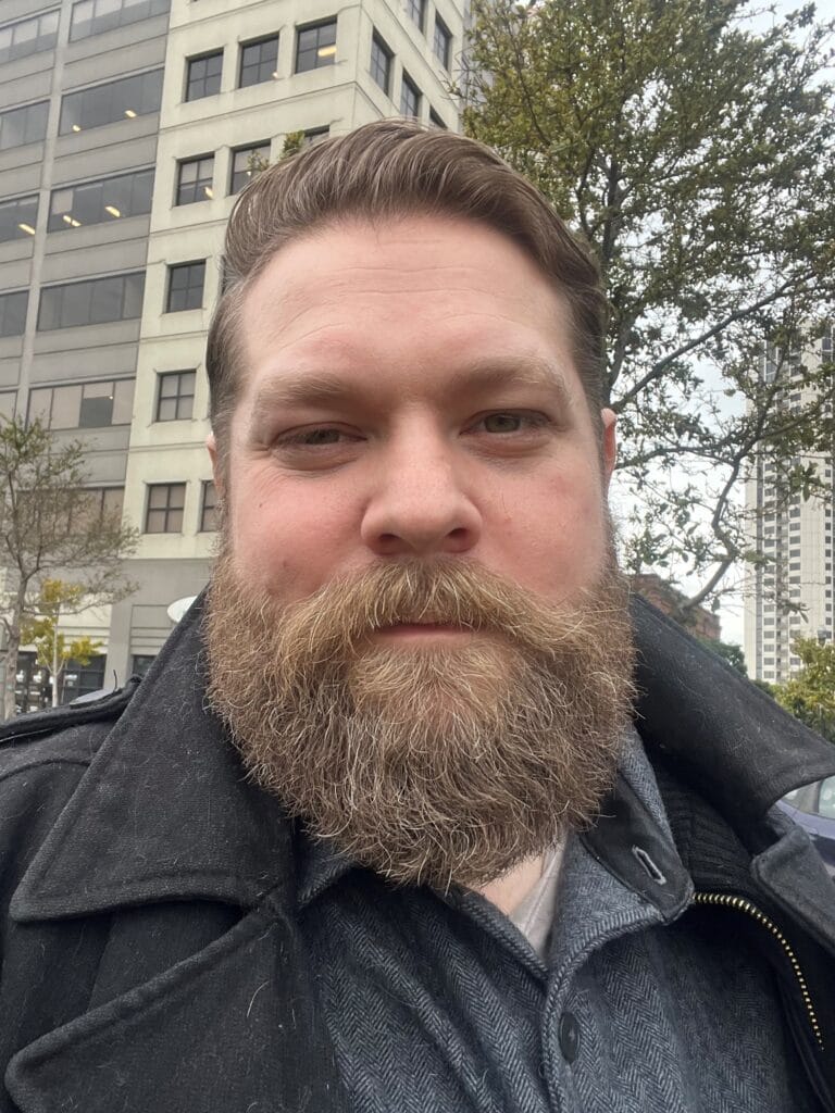 Joel Berger looking into the camera. They are wearing a black coat and have a full beard.