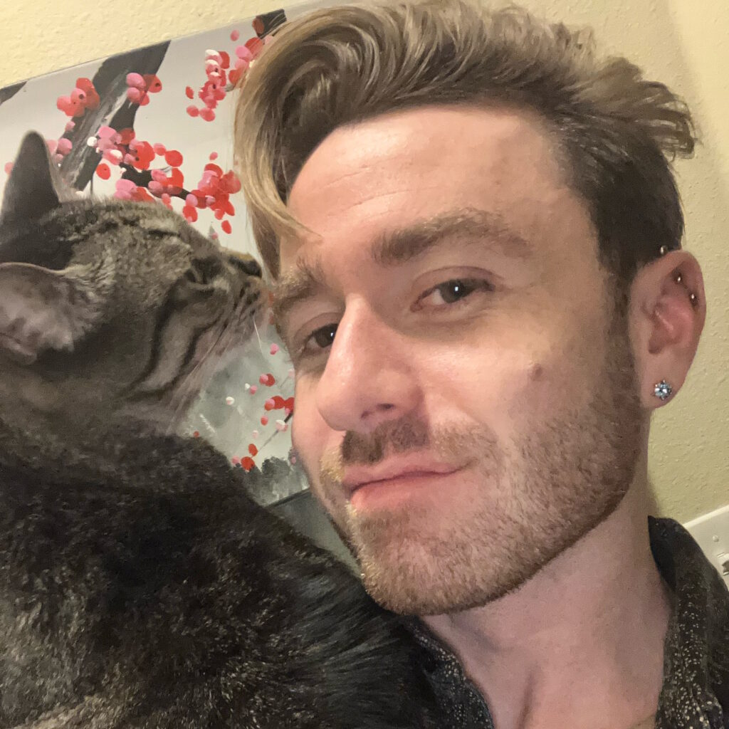 Cameron Kaiser smiling at the camera with a cat next to him, sniffing his face. He has several visible ear piercings.