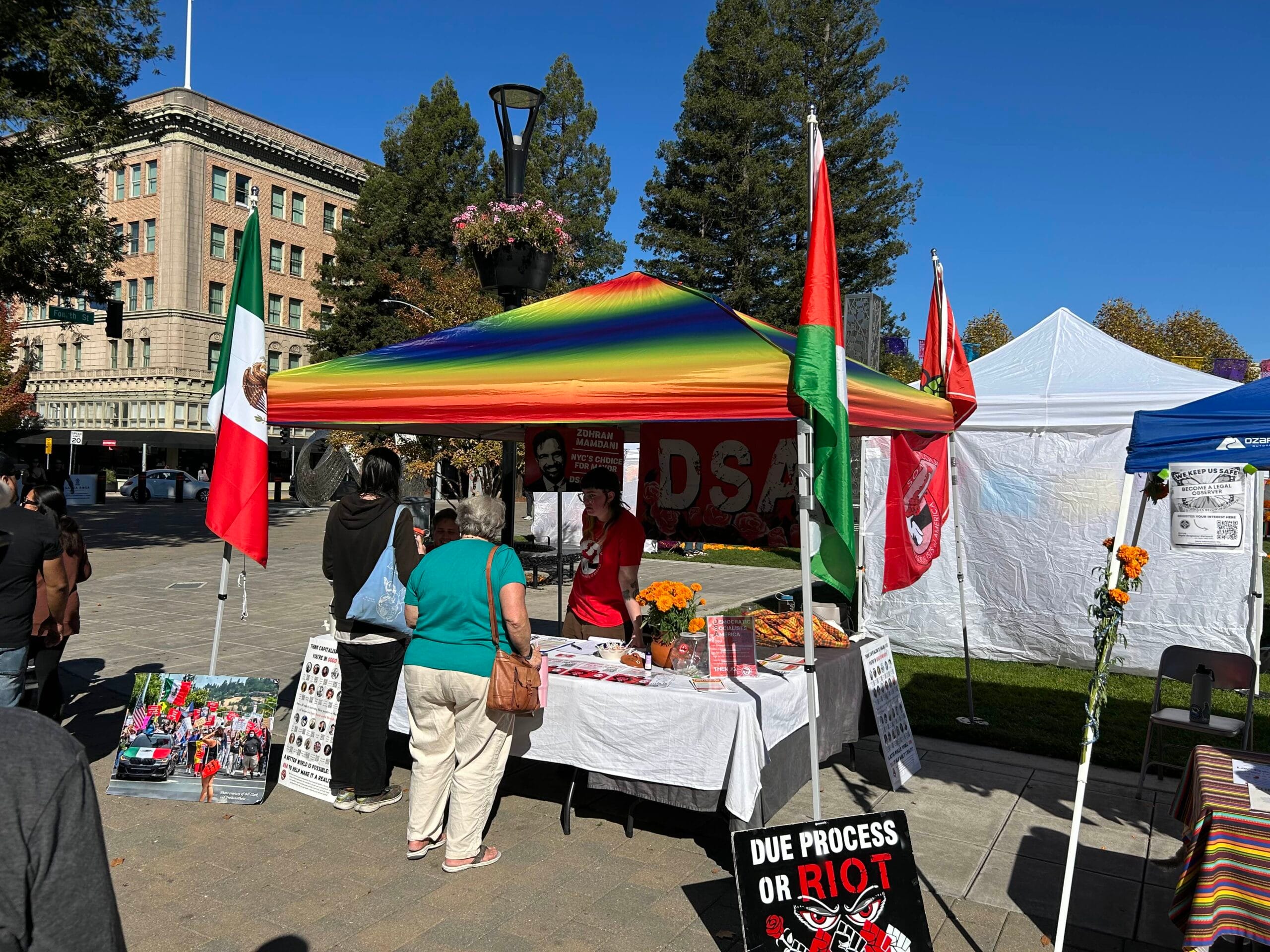 Photo of SoCo DSA's table at Planeta Cosecha's Dia de los Muertos under a rainbow tent featuring Mexican, Palestinian, and DSA flags and a sign that says "Due process or riot."