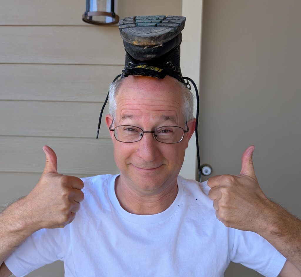 Ian Munc giving a double thumbs-up with a boot upside-down on his head. His white t-shirt matches his hair.