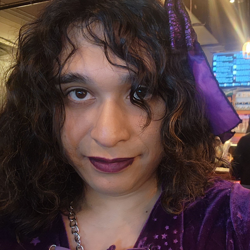 Estel Quintana wearing a purple outfit with matching purple lipstick. Some of her dark curls are falling in front of her face.