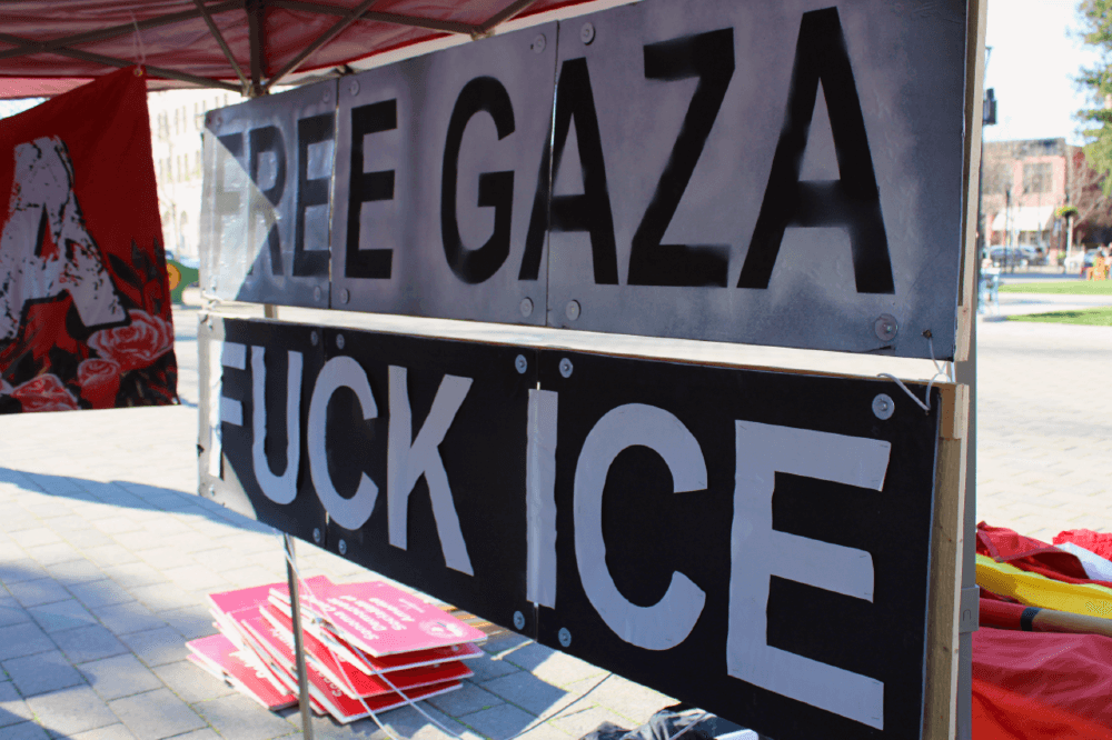 Two large black and white wooden signs reading "Free Gaza" and "Fuck ICE" suspended at the back of our tent in Santa Rosa's Old Courthouse Square, other signs and flags visible in the background.