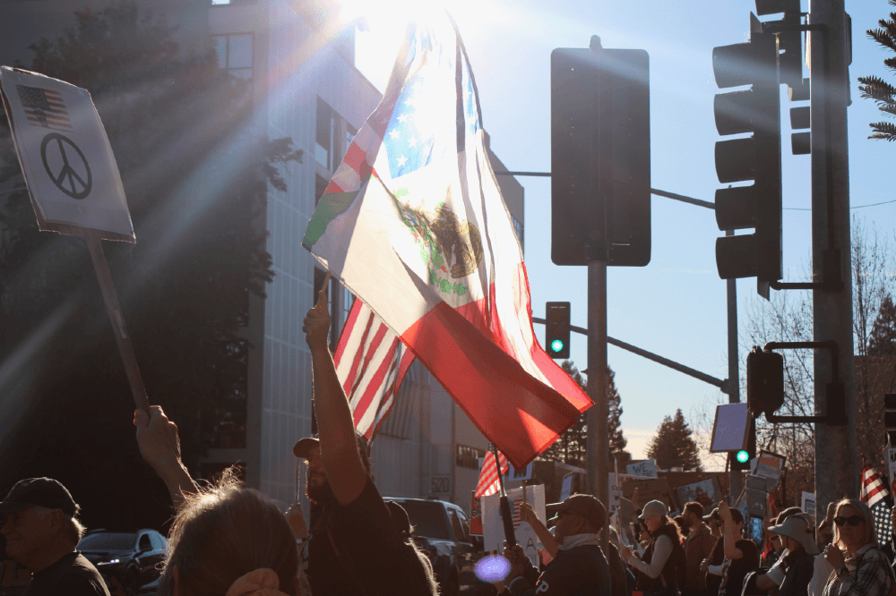Amidst a crowd of protesters, a hand raises the Mexican flag in front of an American flag and a peace picket sign, backlit by the sun in Santa Rosa's Old Courthouse Square.