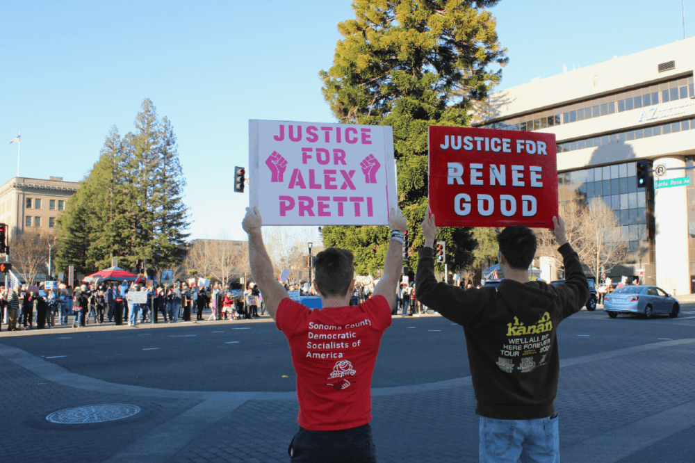 Photo of two of our members from behind, holding signs that read "Justice for Alex Pretti" and "Justice for Renee Good" in downtown Santa Rosa. Across the street in Old Courthouse Square, out of focus, is our tent and a crowd of fellow protesters.
