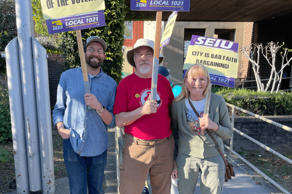 Three of our members hold picket signs on the SEIU 1021 picket line outside Sebastopol City Hall, smiling.