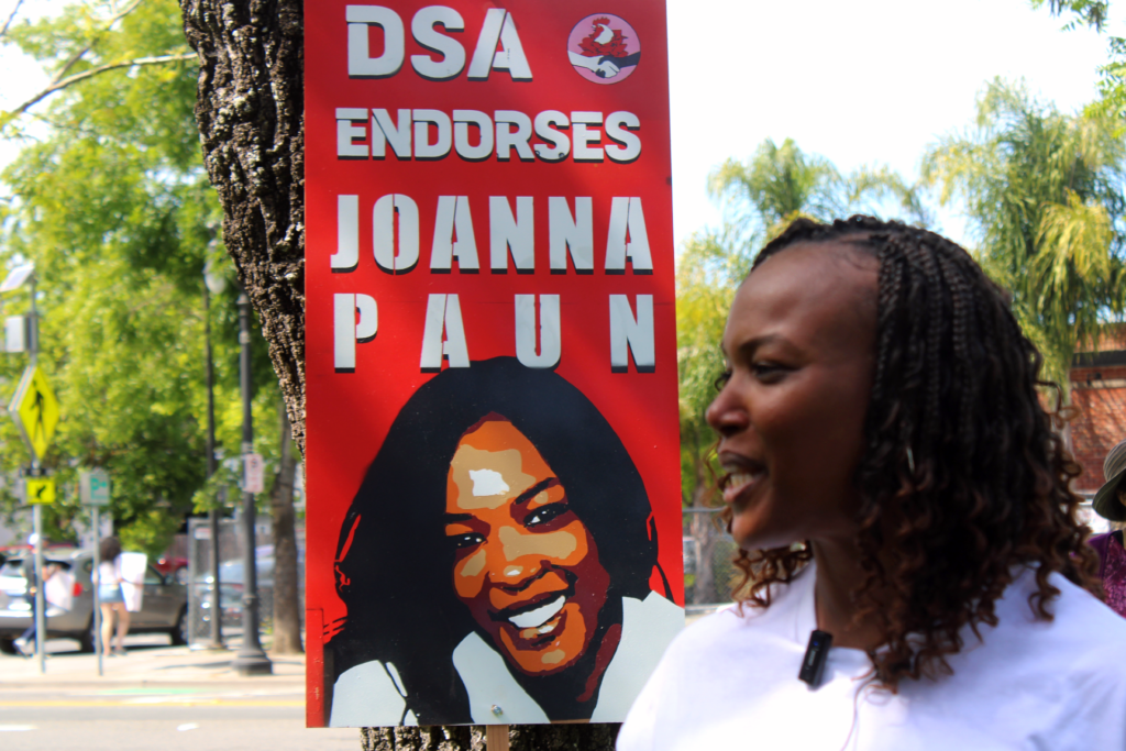 Joanna Paun, a Black woman with short curly hair wearing a white tee shirt, speaks in soft focus in front of a large red sign that reads "DSA Endorses Joanna Paun" in Petaluma's Walnut Park.