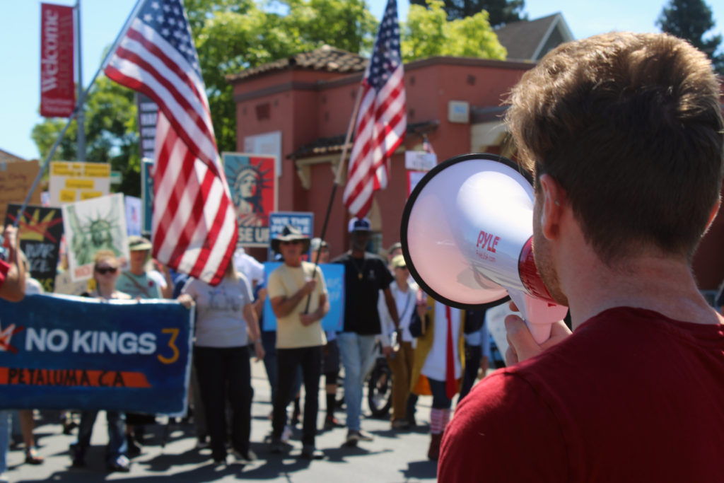 One of our members in a red DSA shirt addresses an out-of-focus crowd with a bullhorn in front of a banner reading "No Kings 3, Petaluma, CA."