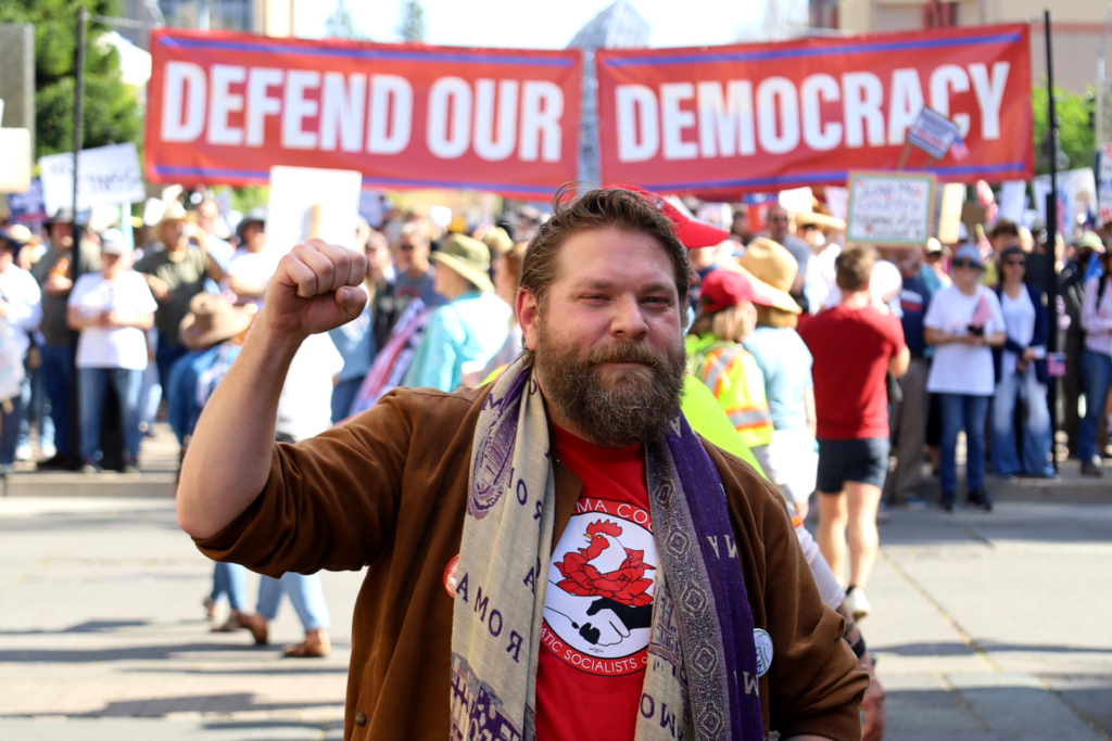 Our Co-Chair, Joel, a white person with brown hair and a full beard wearing a SoCo DSA shirt, holds up a fist in front of a crowd and banner that reads "Defend our Democracy" in front of Santa Rosa's Old Courthouse Square.