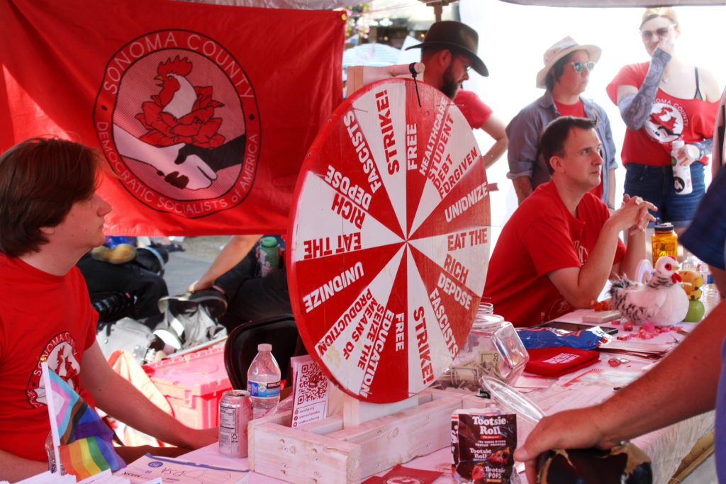 Our members, dressed in red SoCo DSA shirts, sit at one of our tables in Santa Rosa's Old Courthouse Square, SoCo DSA flag behind them, literature, water, and Wheel of Socialism on the table.