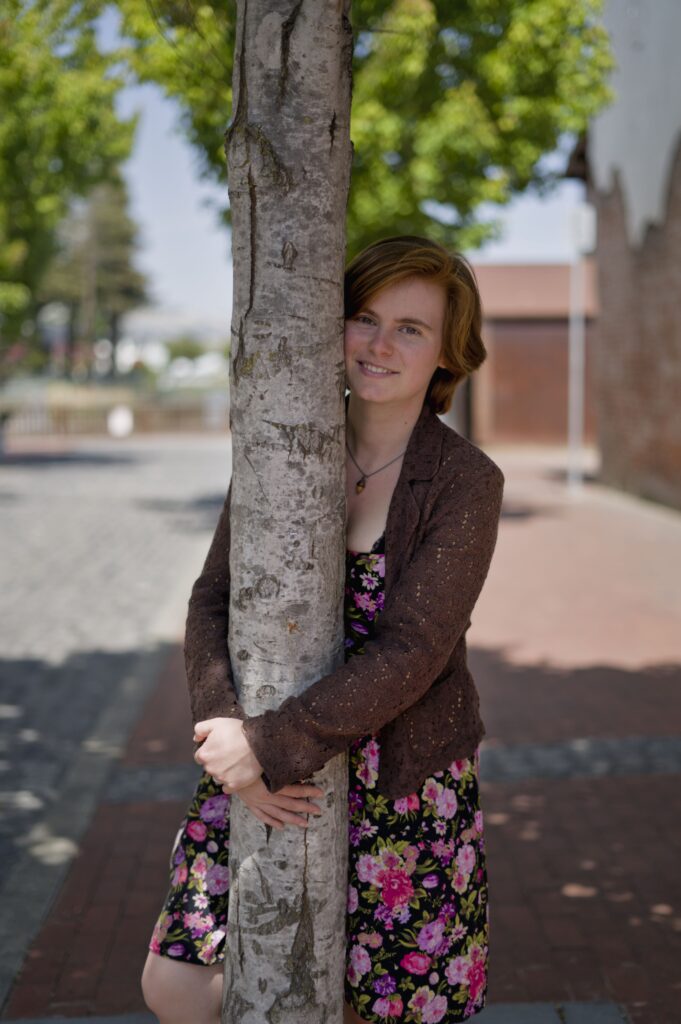 A woman with short auburn hair and fair skin in a floral dress and brown sweater hugs the trunk of a tree on a sunny, brick-paved lane.