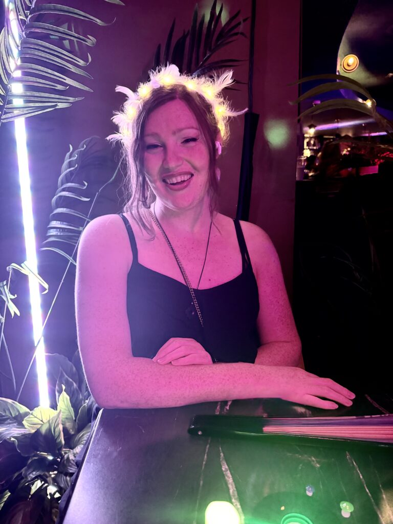 A woman with fair skin and ginger hair smiles in pink light, wearing a black dress.