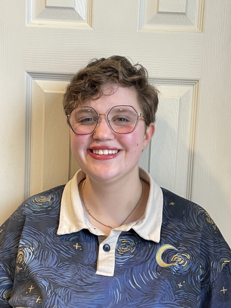 A smiling person with fair skin, short wavy light brown hair, and glasses stands in front of a door, wearing a collared shirt with a print of Vincent Van Gogh's Starry Night.