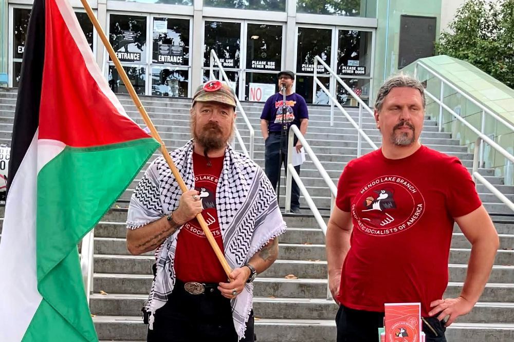 Two members wearing Mendo-Lake Branch shirts stand in front of the steps of Ukiah City Hall, one carrying a Palestinian flag, in front of a speaker on May Day.